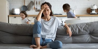 Woman on sofa in living room looking stressed while children play behind her